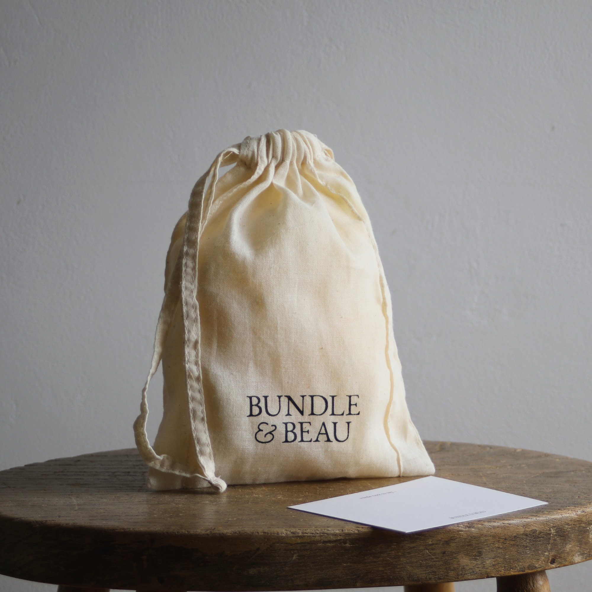 Beige drawstring bag with 'Bundle & Beau' text on a wooden stool against a gray background