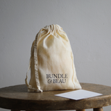 Beige drawstring bag with 'Bundle & Beau' text on a wooden stool against a gray background