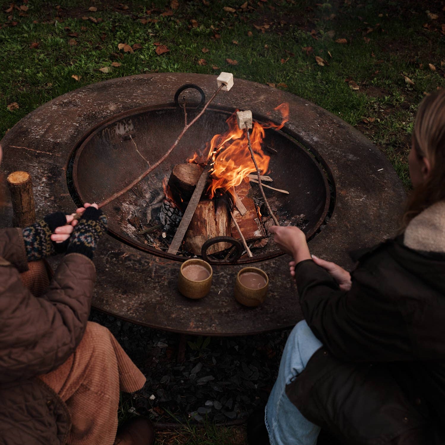 Two people sitting around a round outdoor fire pit, roasting marshmallows and drinking hot chocolate over a small campfire and holding two small brown cups on a grassy lawn at night.