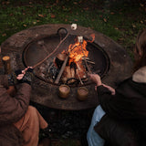 Two people sitting around a round outdoor fire pit, roasting marshmallows and drinking hot chocolate over a small campfire and holding two small brown cups on a grassy lawn at night.