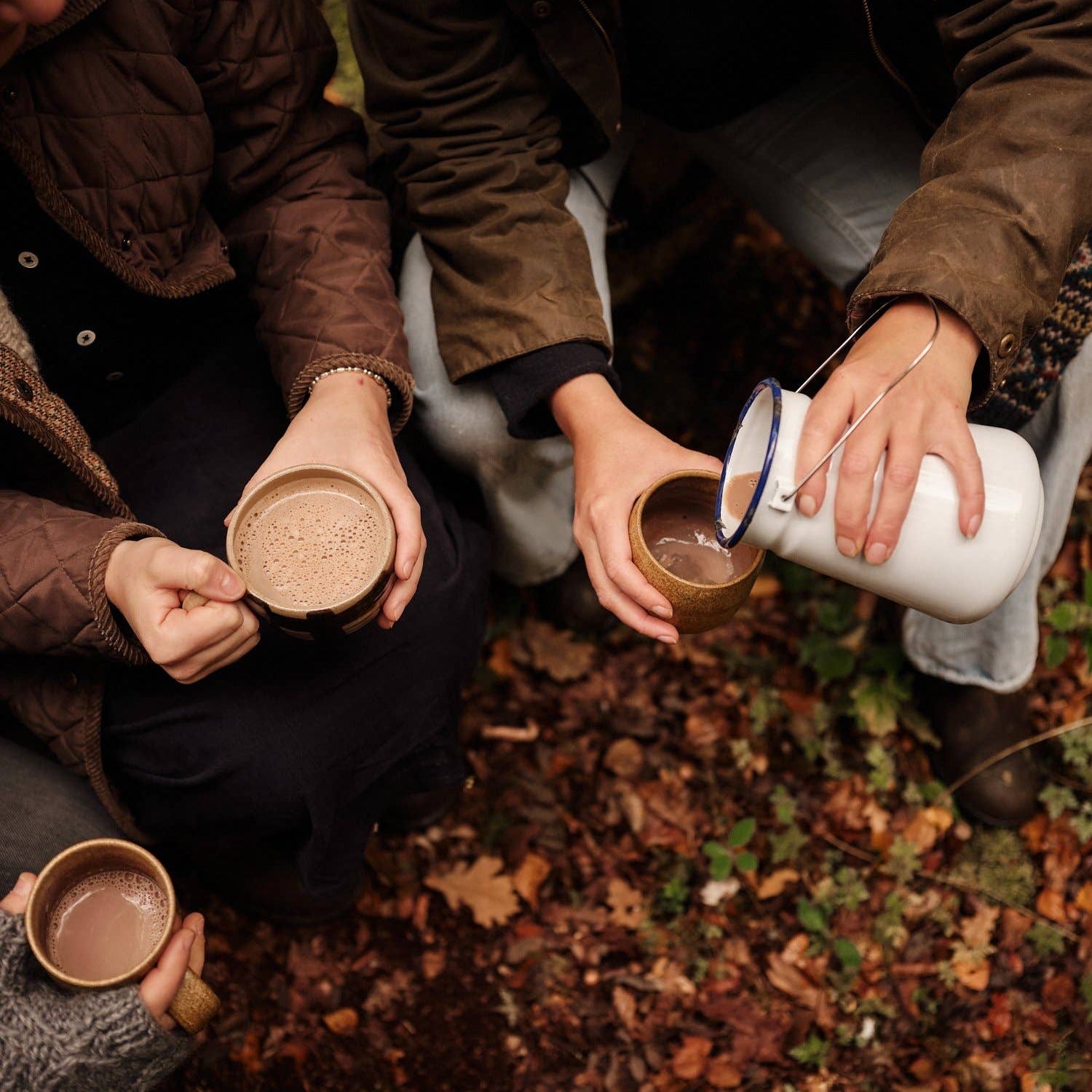 Two people outdoors on a leaf-covered ground pouring hot chocolate from a white enamel pitcher into ceramic cups.