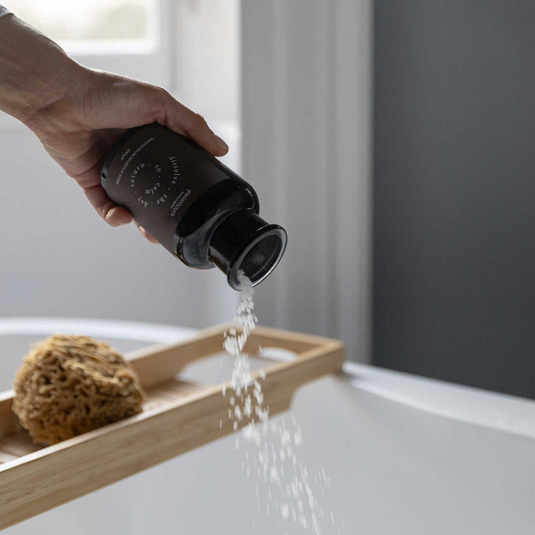 A hand holds a matte black bottle and pours bath salts in the the bath