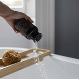 A hand holds a matte black bottle and pours bath salts in the the bath