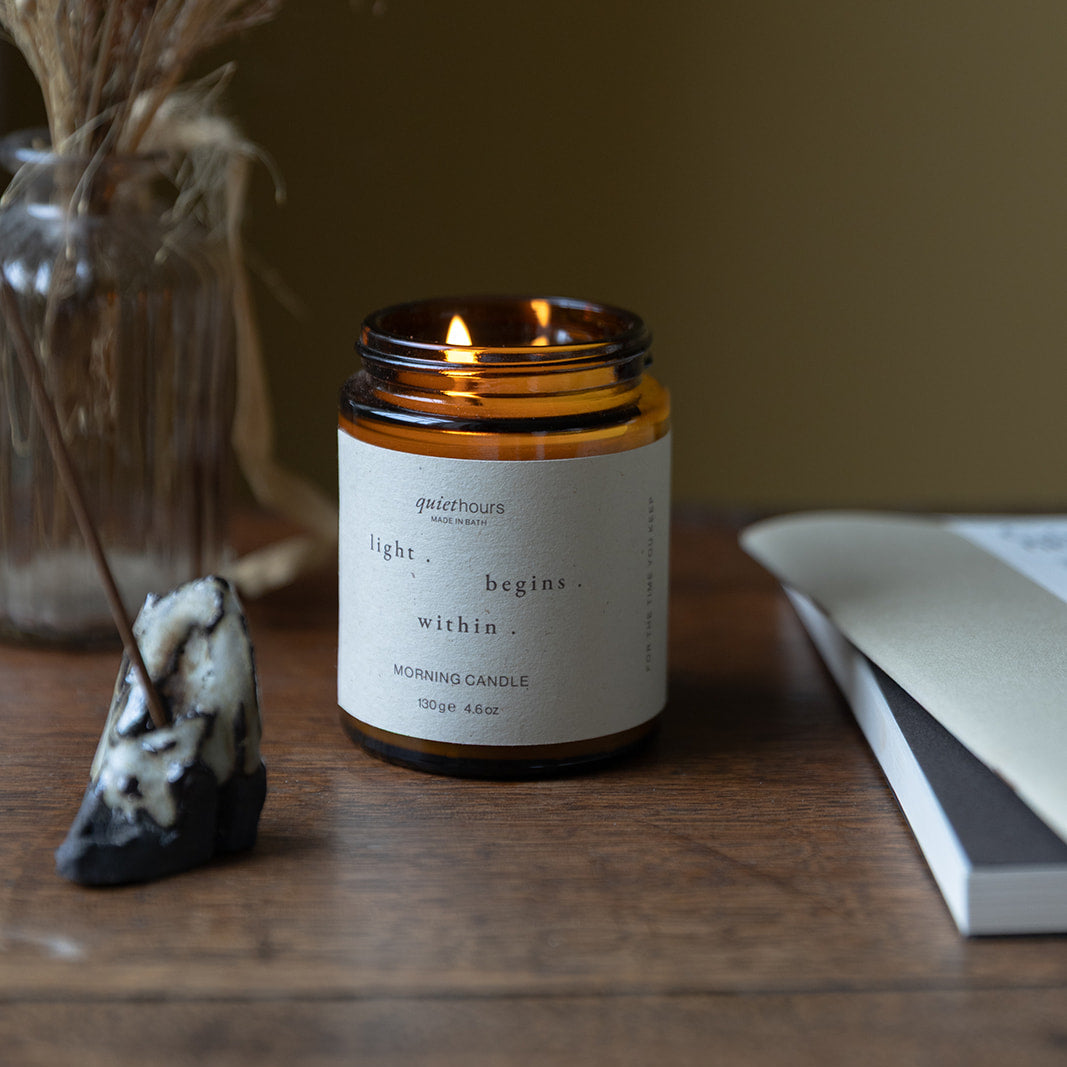 Candle in a amber jar with a white label on a wooden surface, surrounded by decorative items.