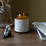 Candle in a amber jar with a white label on a wooden surface, surrounded by decorative items.