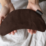 A person holds a curved dark brown fabric eye mask against a soft white sheet background. The mask has a small black brand tag that reads 'paradox'.