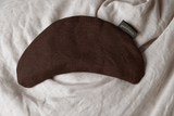 A dark brown curved fabric eye mask or sleep mask laid flat on a textured off-white linen surface. A small black fabric tag with white text is visible near the top right edge of the mask.