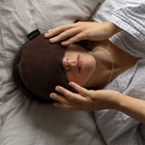 A woman lying in bed wearing a dark brown sleep mask, with her hands resting on the mask and a white button-up shirt.
