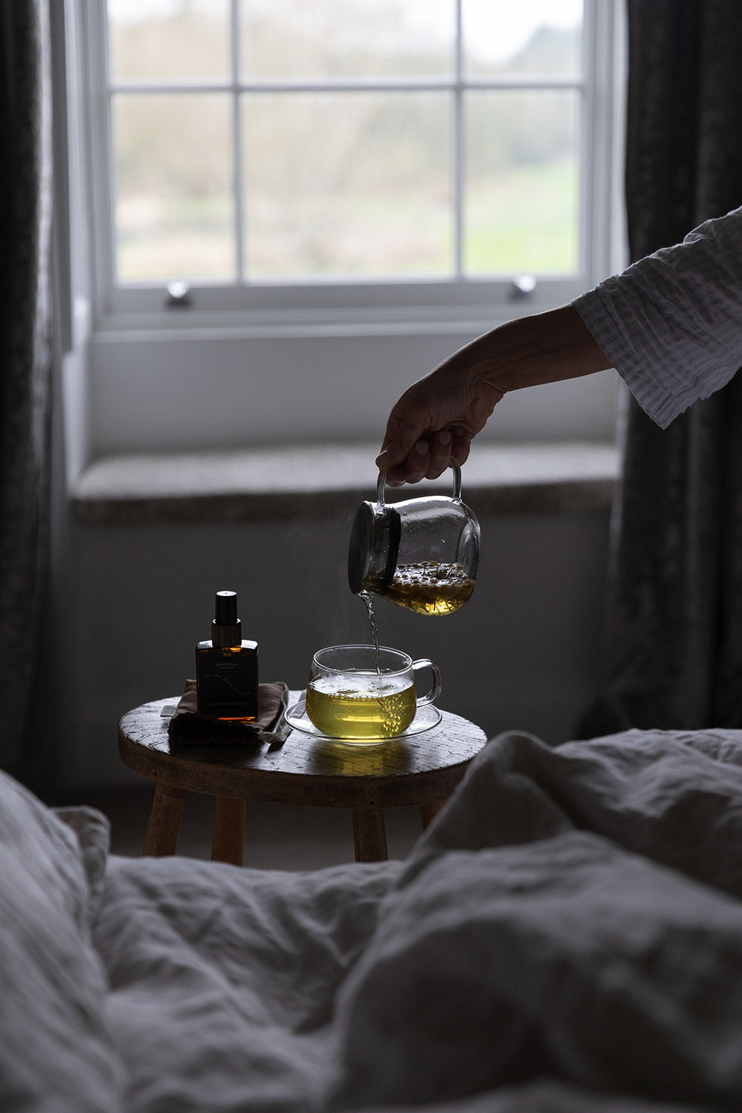 A hand pours tea from a glass teapot into a glass cup on a small wooden table beside a bed, with a window in the background.