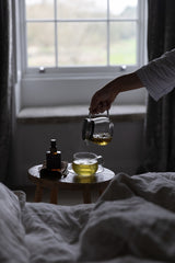 A hand pours tea from a glass teapot into a glass cup on a small wooden table beside a bed, with a window in the background.