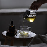 Person pouring tea from a glass teapot into a cup on a wooden table.