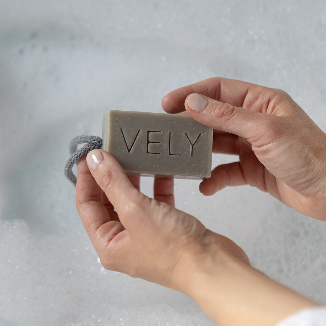 A person's hands holding a rectangular bar of VELY soap in a bubbly, foamy bath setting.