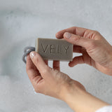 A person's hands holding a rectangular bar of VELY soap in a bubbly, foamy bath setting.