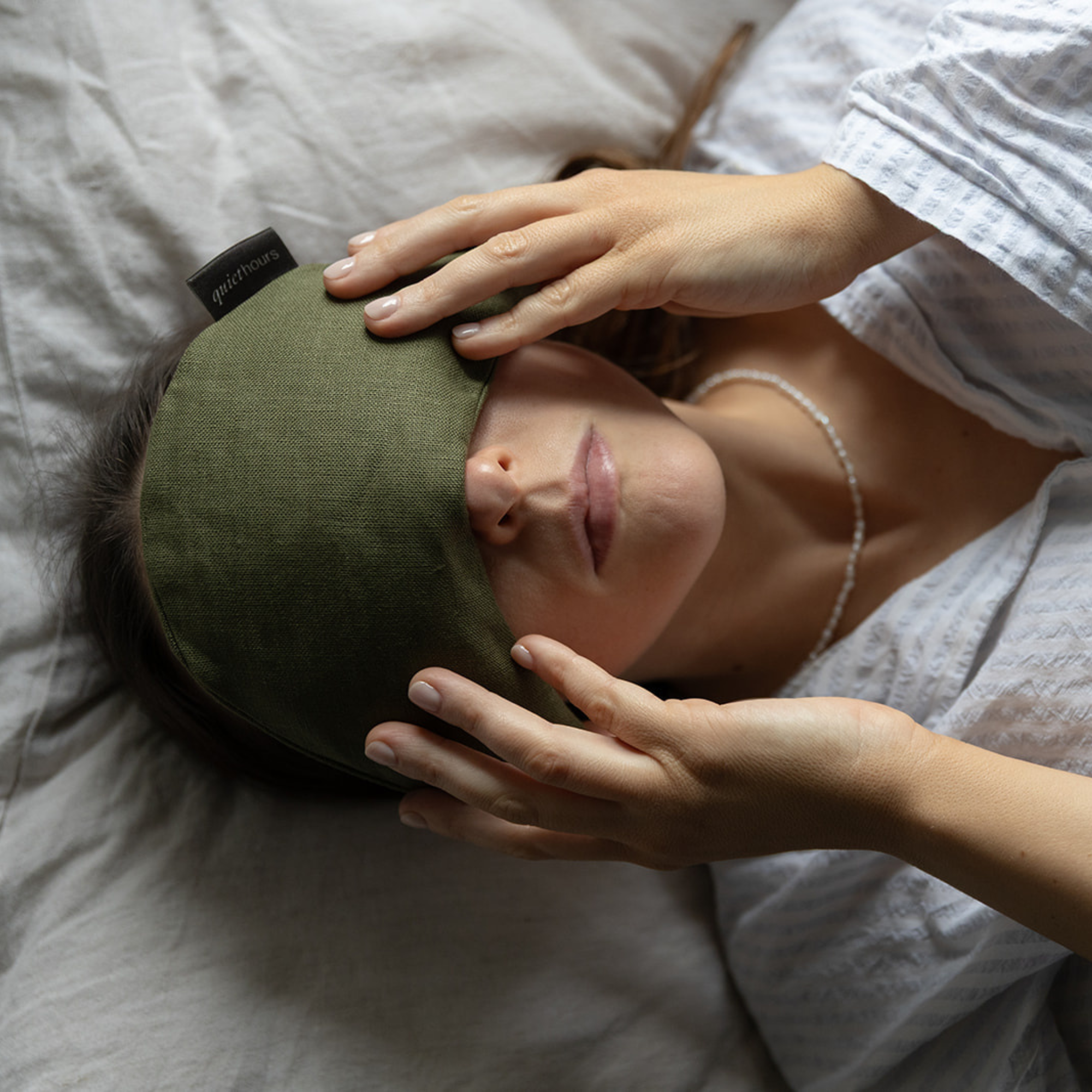 A woman lying in bed with her eyes closed, wearing an olive green weighted eye pillow/ sleep mask. Her hands are gently resting on the mask, and she is wearing a white textured shirt and a delicate pearl necklace.
